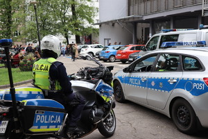 Na zdjęciu policjant w kasku, siedzący na motocyklu służbowym, obok radiowozy służbowe. W oddali zaparkowane na parkingu samochody, oraz ludzie.