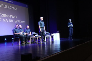 Na zdjęciu pięć osób na scenie. Czterech policjantów siedzących na krzesłach i jedna stojąca policjantka, przemawiająca do mikrofonu. Za nimi wyświetlony napis Debata społeczna, Bezpieczeństwo, możesz mieć na nie wpływ.