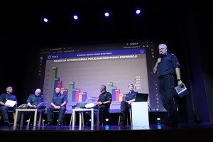 Na zdjęciu pięć osób siedzących na krzesłach, na scenie i jeden stojący, trzymający w ręku mikrofon. Za nimi wyświetlony wykres słupkowy