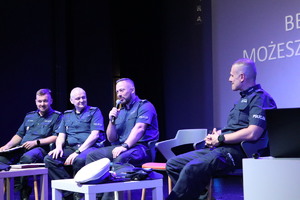 Na zdjęciu czterech siedzących na krzesłach mundurowych na scenie, jeden z nich trzymający w ręku mikrofon.