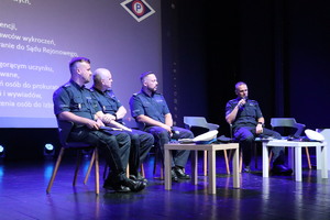 Na zdjęciu, na cenie czterech siedzących policjantów, jeden z nich przemawia do mikrofonu