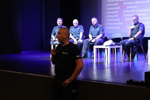 Na zdjęciu przemawiający do mikrofonu policjant stojący przed sceną, za nim na scenie czterech funkcjonariuszy policji