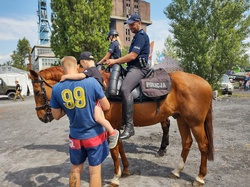 Na zdjęciu dwa konie służbowe, na nich policjant i policjantka obok mężczyzna z kilkuletnim chłopcem na rękach.