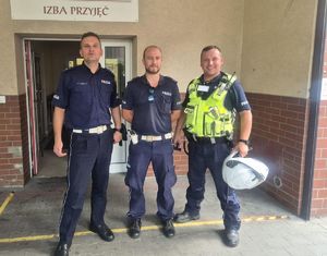 Na zdjęciu trzech uśmiechniętych policjantów stojących przed budynkiem szpitala.