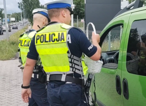Na zdjęciu widzimy dwóch policjantów, stojących przy samochodzie zielonego koloru. Jeden z nich podaje koszyk do okna kierowcy.