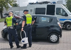 Na zdjęciu dwóch policjantów przy samochodzie osobowym ciemnego koloru, jeden trzyma koszyk. Za nimi radiowóz policyjny.