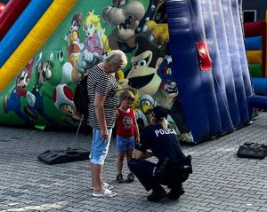 Na zdjęciu policjantka rozmawiająca z kilkuletnim chłopcem trzymanym za rękę przez kobietę, obok nich dmuchana zjeżdżalnia dla dzieci.