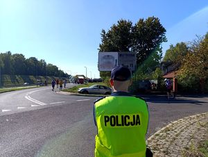 Na zdjęciu policjant w żółtej kamizelce odblaskowej, przed nim biegnące w półmaratonie osoby.