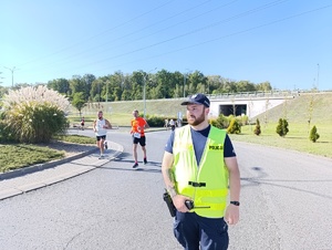 Na zdjęciu policjant w żółtej kamizelce odblaskowej, obok niego i za nim biegnące w półmaratonie osoby.