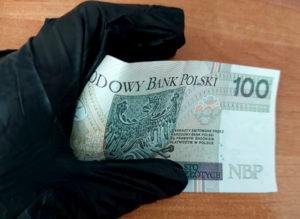 na zdjęciu dłoń w czarnej rękawiczce trzymająca banknot 100 złotych