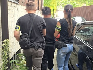 na zdjęciu zatrzymany mężczyzna ubrany na czarno, prowadzony przez nieumundurowanego policjanta i policjantkę.