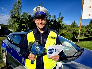 na zdjęciu przed radiowozem policjant ruchu drogowego, trzyma w dłoni ulotki mistrzu zwolnij, w drugiej dłoni ma urządzenie do pomiaru prędkości