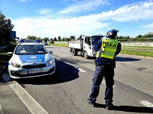 na zdjęciu obok radiowozu policjant stojący na drodze i wykonujący pomiar prędkości