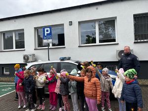 na zdjęciu dzieci przed radiowozem, trzymają w dłoniach odblaski, obok stoi policjant, wszyscy przed komisariatem