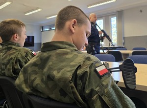 na zdjęciu licealiści w strojach moro siedzą przy stołach, oglądają broń służbową, przy nich policjant