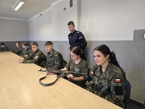 na zdjęciu licealiści w strojach moro siedzą przy stołach, oglądają broń służbową, przy nich policjant