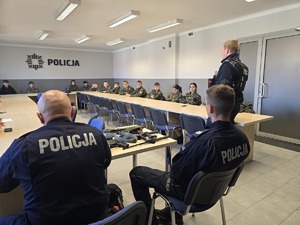 na zdjęciu licealiści w strojach moro siedzą przy stołach, przed nimi policjant ,obok broń służbowa i dwóch policjantów