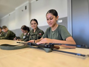 na zdjęciu licealiści w strojach moro siedzą przy stołach, oglądają broń służbową