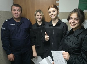 na zdjęciu trzy dziewczyny w czarnych moro trzymają kartki z odciskami palców, obok policjant,