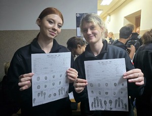 na zdjęciu dwie uśmiechnięte licealistki, trzymają w dłoniach kartki na których są wykonane ich odciski palców, pozują do zdjęcia