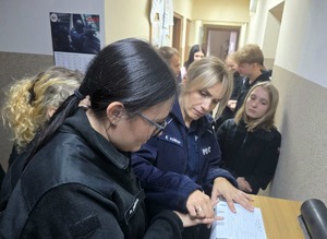 na zdjęciu policjantka przykłada dłoń licealistki do kartki papieru by wykonać odciski palców, przy niej licealiści w czarnych strojach