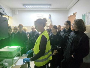 na zdjęciu techniczka kryminalistyki przy urządzeniu do daktyloskopii w żółtej kamizelce, które świeci za zielono, przy niej licealiści w czarnych strojach