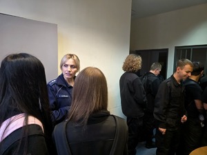na zdjęciu policjantka, obok licealiści przy kratach, w pomieszczeniu dla zatrzymanych osób