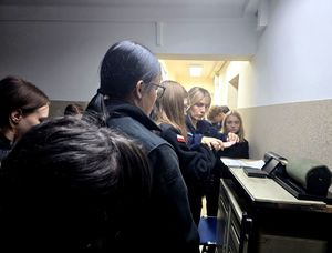 na zdjęciu policjantka przykłada dłoń licealistki do kartki papieru by wykonać odciski palców, przy niej licealiści w czarnych strojach