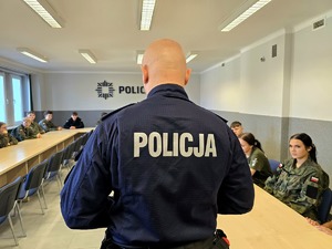 na zdjęciu policjant stojący przed uczniami siedzącymi przy stołach, są w strojach moro