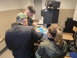 na zdjęciu zatrzymany mężczyzna, obok nieumundurowana policjantka, przed nimi policyjny technik w trakcie pobierania odcisków palców