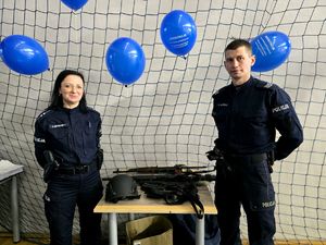 na zdjęciu na sali policjant, policjantka i broń na stoliku, za nimi niebieskie balony
