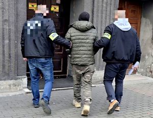 na zdjęciu zatrzymany w kajdankach zespolonych prowadzony przez dwóch nieumundurowanych policjantów, przed wejściem do prokuratury