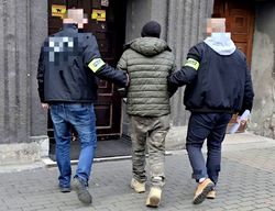 na zdjęciu zatrzymany w kajdankach zespolonych prowadzony przez dwóch nieumundurowanych policjantów, przed wejściem do prokuratury