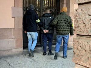 na zdjęciu przed budynkiem sądu, dwóch nieumundurowanych policjantów prowadzących zatrzymanego w kajdankach