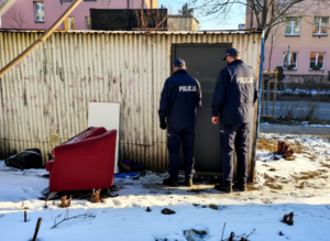 na zdjęciu dwaj policjanci przed budynkiem przypominającym garaż, przed którym stoi stary, czerwony fotel