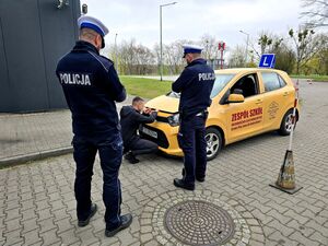 widzimy dwóch policjantów drogówki, przed nimi chłopak przy samochodzie do jazdy egzaminacyjnej podnoszący pokrywę silnika pojazdu