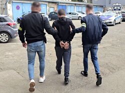 na parkingu policyjnym dwóch nieumundurowanych policjantów prowadzi zatrzymanego mężczyznę, zatrzymany ma dłonie w kajdankach zapiętych z tyłu