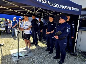 młody mężczyzna, syn Marka Sienickiego stojący pod namiotem i przemawiający przed mikrofonem, za nim mundurowi