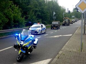 motocykl służbowy stojący na jezdni, za nim radiowóz policyjny i kolumna pojazdów militarnych
