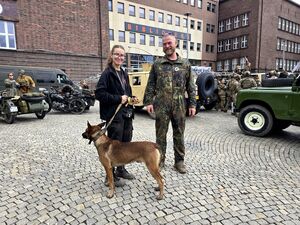 mężczyzna i kobieta, która trzyma psa, dookoła nich pojazdy militarne i zgromadzone osoby
