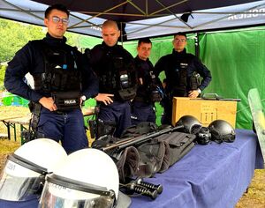 czterech policjantów stoi przy policyjnym stoisku, ma stole leży sprzęt służbowy kaski, kamizelki, broń, przy nich tarcze