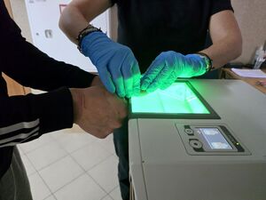 pobieranie odcisków palców, dłonie policjantki w niebieskich rękawiczkach na kciukach zatrzymanego mężczyzny przyłożonych do urządzenia daktyloskopijnego