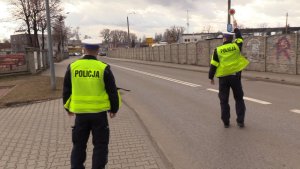 Na pierwszym planie widocznych jest dwóch policjantów, jeden stoi na chodniku, drugi na jezdni przy użyciu tarczy zatrzymuje samochód dostawczy