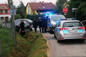 grafika-dzień, policyjny radiowóz z włączonymi sygnałami uprzywilejowania, uszkodzony skradziony VW Golf w rowie, pomiędzy pojazdami policjanci