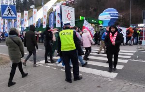 grafika-dzień, policjant stoi przy drodze, kieruje ruchem, w tle skocznia