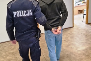 zdjęcie-pomieszczenie, oświetlenie naturalne i sztuczne, policjant prowadzi trzymając za przedramię, mężczyznę, który ma kajdanki na rekach trzymanych z tyłu