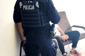 policjant w mundurze pilnuje zatrzymanego mężczyzny. Zatrzymany ma kajdanki założone na ręce i nogi