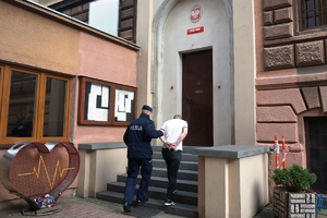 zdjęcie- dzień, pochmurno, policjant prowadzi mężczyznę po schodach do zakładu karnego, mężczyzna ma kajdanki na rękach założonych z tyłu