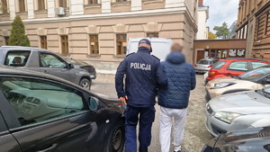 zdjęcie- dzień, policjant prowadzi zatrzymanego mężczyznę, trzyma go za ramię, mężczyzna ma kajdanki na rękach z przodu oraz nogach w rejonie kostek