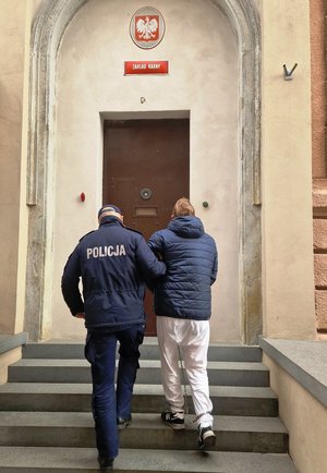 zdjęcie- dzień, policjant prowadzi zatrzymanego mężczyznę, trzyma go za ramię, mężczyzna ma kajdanki na rękach z przodu oraz nogach w rejonie kostek